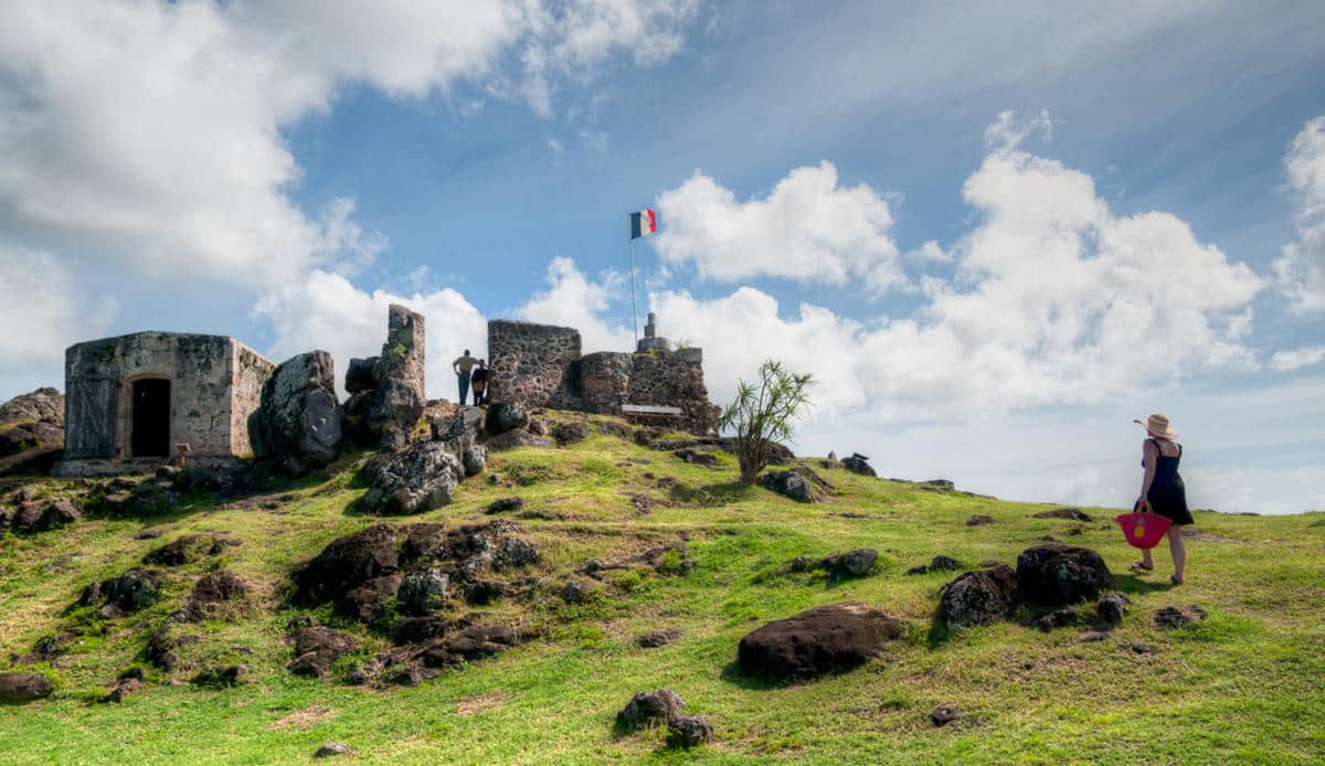 Ruined stone fortress on a grassy hill with a small flag atop, visitors near the ruins, and a woman in a hat walking with a pink bag under a blue sky.